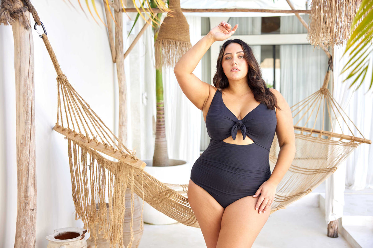 A curvy woman from Chicago is relaxing in an overwater bungalow in the Maldives, wearing the Aria Black Cezanne plus size underwire cutout one piece, selected for its exceptional lift for sagging breasts, luxury tummy control shaping, high end designer swimsuit look, and premium quality curvy swim fit. She bought this swimsuit from Sun Vixen Swimwear because it gives her a sexy elegant resort look perfect for photos, private pools, and lagoon lounging. Women across Canada and the USA buy this for vacations