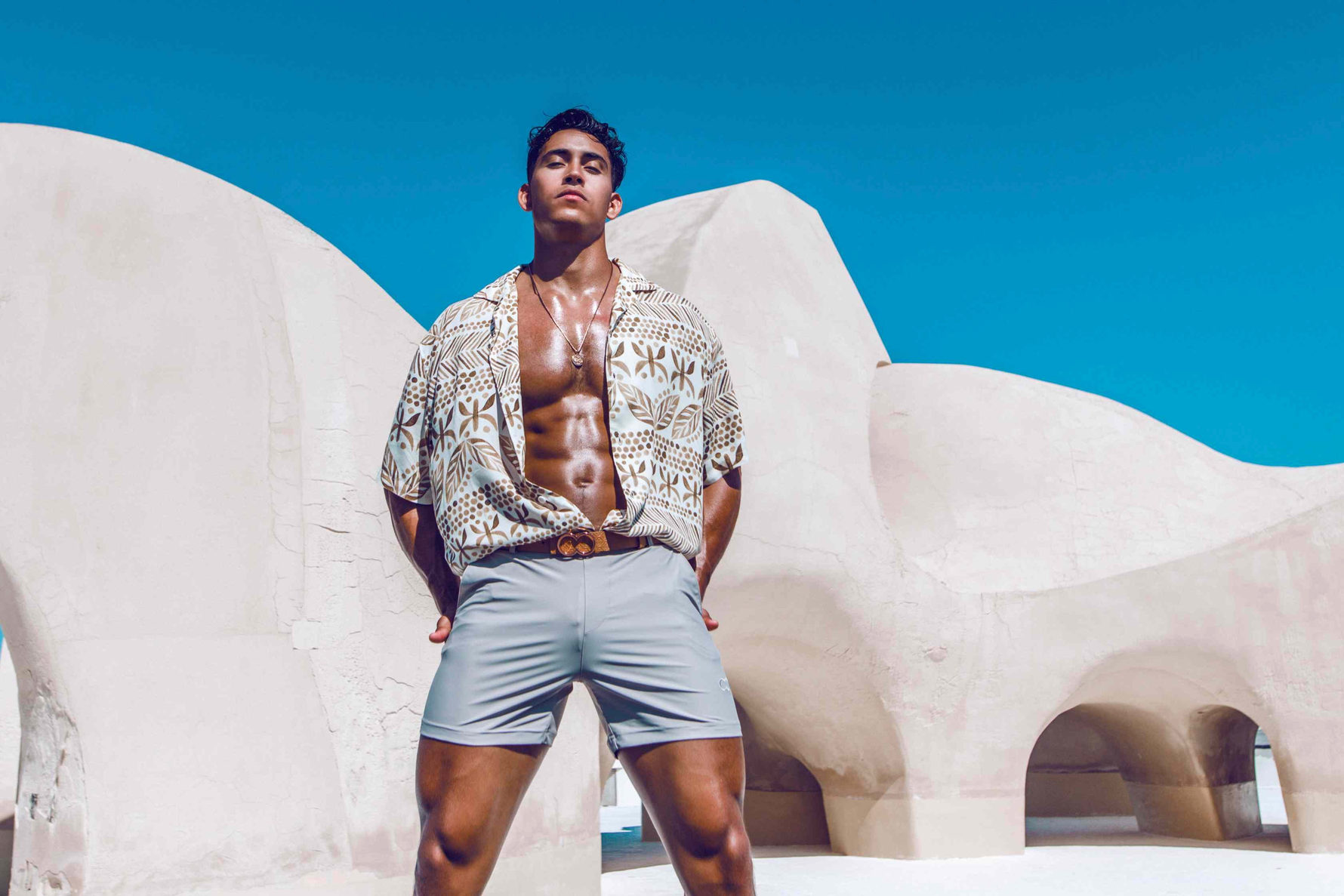 A hot guy wears stylish short swim trunks from Sun Vixen Swimwear on his resort vacation, showcasing sleek grey swim trunks that complement his physique. These fitted swim trunks, featuring a slim fit design and 4 inch inseam, are among the best men's swim trunks look. This mens designer swimwear store sell mens short swim trunks and euro style swim shorts. The collection includes slim fit swim trunks, tight swim shorts, sustainable mens beachwear, hottest swim trunks & stylish Hawaiian beach shirts online