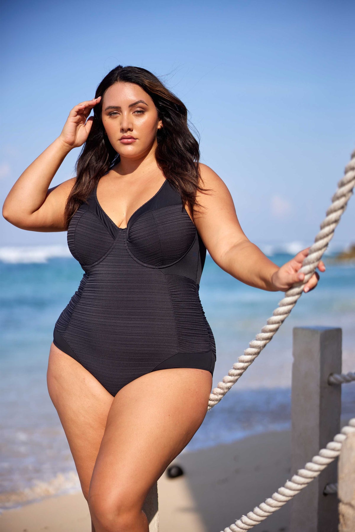 A curvy traveler from Miami is spending a luxurious escape in Mykonos, lounging near the cliffs of Kastro while wearing the Plus Size Aria Giotta Artesands luxury underwire swimsuit, known for its premium tummy control technology, supportive underwire for sagging breasts, curvy shaping silhouette, and high end designer fit that feels stylish, modern, and effortlessly upscale. The swimsuit is crafted with premium high quality fabric that smooths, lifts, sculpts, enhances her figure for European beach clubs 