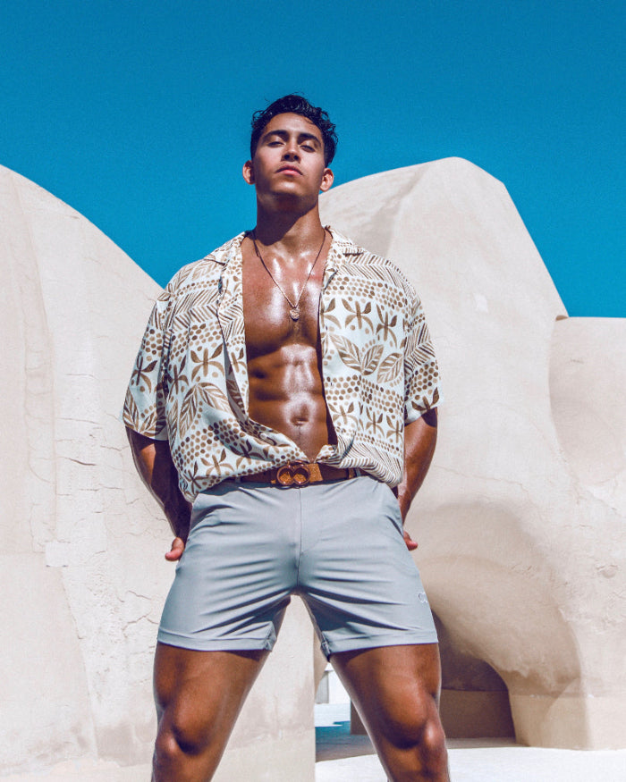 A hot guy wears stylish short swim trunks from Sun Vixen Swimwear on his resort vacation, showcasing sleek grey swim trunks that complement his physique. These fitted swim trunks, featuring a slim fit design and 4 inch inseam, are among the best men's swim trunks look. This mens designer swimwear store sell mens short swim trunks and euro style swim shorts. The collection includes slim fit swim trunks, tight swim shorts, sustainable mens beachwear, hottest swim trunks & stylish Hawaiian beach shirts online